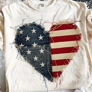 Op de afbeelding: Crèmekleurig T-shirt met een hartvormig ontwerp. De linkerkant van het hart is van denim met witte sterren, en de rechterkant heeft rood-witte strepen, die lijken op de Amerikaanse vlag. De randen zijn gerafeld.