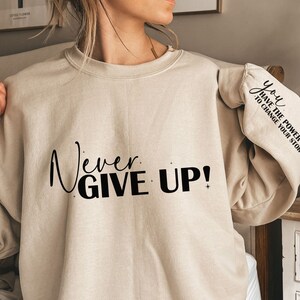 Op de afbeelding: Een beige sweatshirt met de tekst "Never Give Up!" gedrukt op de voorkant en "You Have The Power To Change Your Story" gedrukt op de mouw.