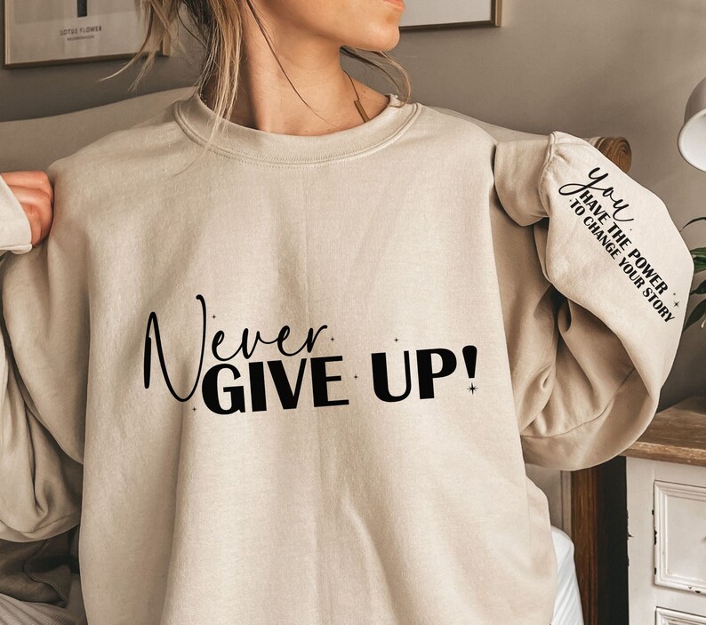 Op de afbeelding: Een beige sweatshirt met de tekst "Never Give Up!" gedrukt op de voorkant en "You Have The Power To Change Your Story" gedrukt op de mouw.