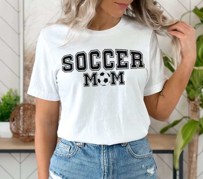 Puede incluir: Camiseta blanca con texto negro que dice "Soccer Mom" y un gr&aacute;fico de bal&oacute;n de f&uacute;tbol blanco y negro.