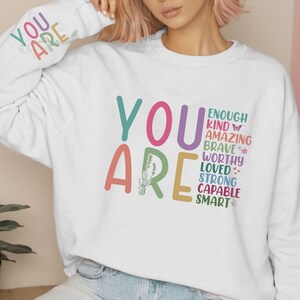 Może przedstawiać: Biała bluza z nadrukiem "YOU ARE" dużymi, kolorowymi literami. Tekst jest ułożony pionowo i zawiera słowa "enough, kind, amazing, brave, worthy, loved, strong, capable, smart".
