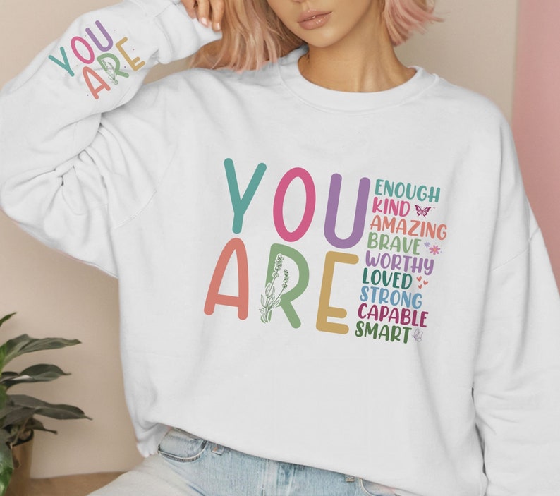 Może przedstawiać: Biała bluza z nadrukiem "YOU ARE" dużymi, kolorowymi literami. Tekst jest ułożony pionowo i zawiera słowa "enough, kind, amazing, brave, worthy, loved, strong, capable, smart".