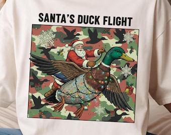 May include: Cream-colored sweatshirt featuring a graphic of Santa Claus riding a duck adorned with Christmas lights. The duck is set against a camouflage background with snowflakes and gift boxes. The text "SANTA'S DUCK FLIGHT" is printed above the graphic.