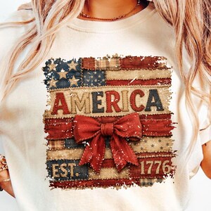May include: A cream-coloured t-shirt with a distressed American flag design. The word "AMERICA" is in bold letters, with a red bow beneath. The year "1776" is printed at the bottom. The shirt has a crew neck. The design is reminiscent of the American flag.