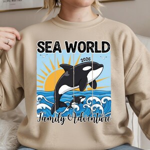K&ouml;nnte beinhalten: Beiges Sweatshirt mit einer Grafik eines schwarz-wei&szlig;en Orcas, der aus blauen Wellen springt, mit einer gelben Sonne und dem Text "SEA WORLD 2026 Family Adventure". Das Design umfasst zwei Orcas und M&ouml;wen.