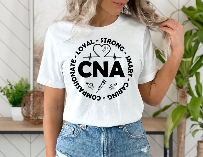 May include: White t-shirt with a black circle graphic that says "CNA" and includes a heart with a heartbeat line, a syringe, and pills. The text around the circle says "Caring - Smart - Strong - Loyal - Compassionate".