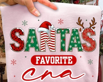 Może przedstawiać: Jasnoróżowa bluza z napisem "SANTA'S FAVORITE CNA" w świątecznym designie. Słowo "SANTA'S" jest w kolorze czerwonym i zielonym, z czapką Świętego Mikołaja. Płatki śniegu i rogi renifera dopełniają świąteczny motyw.