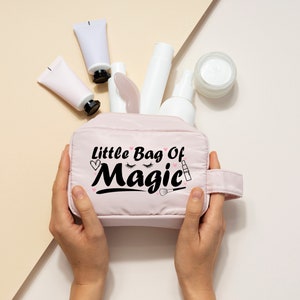 May include: A pink zippered makeup bag with the text "Little Bag Of Magic" in black lettering. The bag is decorated with a heart, eyelashes, and a lipstick. The bag is being held by a person's hands.
