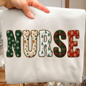Puede incluir: Una sudadera color crema con la palabra "NURSE" en letras grandes y festivas. Cada letra presenta un patrón navideño diferente, que incluye copos de nieve, hombres de jengibre, árboles de Navidad y cuadros escoceses. La sudadera es sostenida por una mano.