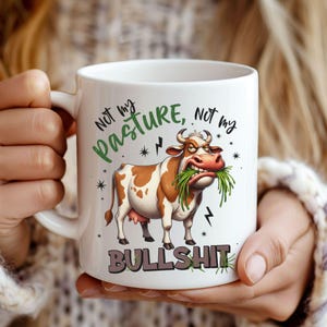 May include: White ceramic mug featuring a cartoon cow with brown and white spots, eating grass. The mug has the text "Not my pasture, not my bullshit" in green and gray. Lightning bolt and star graphics are also present.