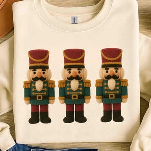 May include: Cream-colored sweatshirt featuring three festive nutcracker soldier designs. Each nutcracker has a red and gold hat, a green and gold uniform, and a black mustache. The design is centered on the front of the sweatshirt.