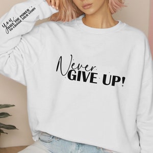 Op de afbeelding: Witte trui met de tekst "Never Give Up!" gedrukt op de voorkant. De linkermouw heeft de tekst "You have the power to change your story" erop gedrukt.