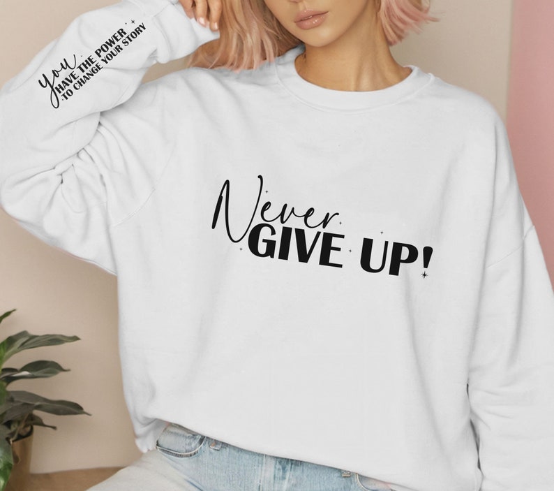 Op de afbeelding: Witte trui met de tekst "Never Give Up!" gedrukt op de voorkant. De linkermouw heeft de tekst "You have the power to change your story" erop gedrukt.
