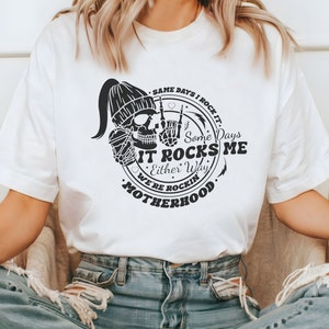 May include: White t-shirt with a black and white graphic design. The graphic features a skull wearing a beanie, holding a coffee cup, and making a rock and roll hand gesture. The text reads "Same days I rock it. Some days it rocks me. Either way we're rockin' motherhood."