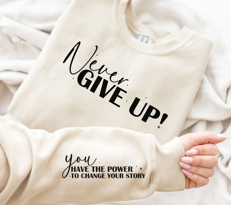 Op de afbeelding: Een cr&egrave;mekleurig sweatshirt met de tekst "Never Give Up!" op de voorkant en "You Have The Power To Change Your Story" op de mouw.