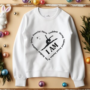 May include: White crew neck sweatshirt with a black heart design. The graphic includes a gymnast silhouette and the words "I AM." The heart is encircled by words such as "Resilient," "Brave," and "Fearless."