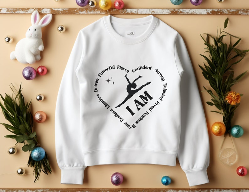 May include: White crew neck sweatshirt with a black heart design. The graphic includes a gymnast silhouette and the words "I AM." The heart is encircled by words such as "Resilient," "Brave," and "Fearless."
