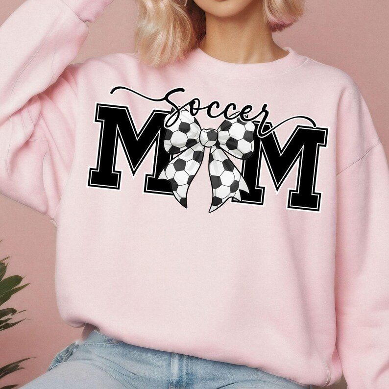 May include: A light pink sweatshirt with the words "Soccer Mom" in black and white. The word "MOM" is in bold, block letters, with a soccer ball bow in the center. The word "Soccer" is written in cursive above the bow.
