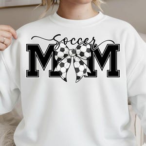 May include: White sweatshirt with a black and white graphic. The graphic says "Soccer Mom" with the letters "MOM" in bold, black block letters. A bow made of soccer balls is in the center of the letters.