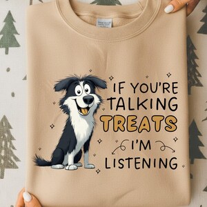 May include: A tan sweatshirt featuring a cartoon dog with black and white fur. The dog has a happy expression and the text "IF YOU'RE TALKING TREATS I'M LISTENING" is printed on the front.
