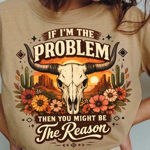 May include: Tan t-shirt featuring a graphic with a cow skull, desert landscape, and flowers. The text reads "IF I'M THE PROBLEM THEN YOU MIGHT BE The Reason."