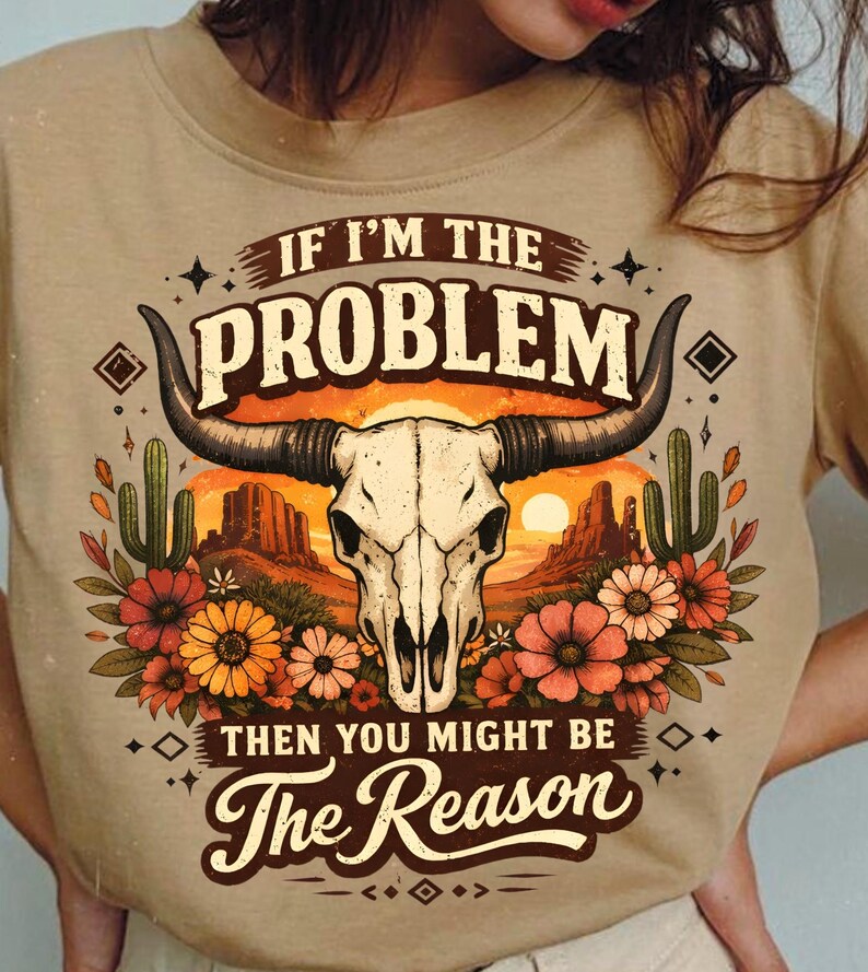 May include: Tan t-shirt featuring a graphic with a cow skull, desert landscape, and flowers. The text reads "IF I'M THE PROBLEM THEN YOU MIGHT BE The Reason."