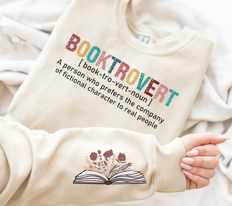 K&ouml;nnte beinhalten: Ein beiger Pullover mit dem Text "BOOKTROVERT [book-tro-vert-noun] A person who prefers the company of fictional character to real people" auf der Vorderseite. Der Pullover hat ein Motiv eines ge&ouml;ffneten Buches mit Blumen auf dem &Auml;rmel.