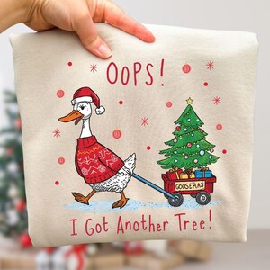 May include: Beige sweatshirt with a whimsical Christmas design. A goose wearing a Santa hat and red sweater pulls a wagon with a decorated Christmas tree. Text reads "OOPS! I Got Another Tree!" and "GOOSEMAS".