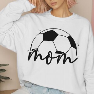 Puede incluir: Sudadera blanca con un gr&aacute;fico de bal&oacute;n de f&uacute;tbol negro y la palabra "mom" en letra cursiva.