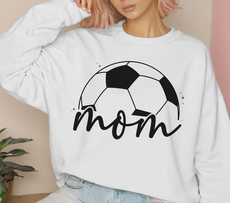 Puede incluir: Sudadera blanca con un gr&aacute;fico de bal&oacute;n de f&uacute;tbol negro y la palabra "mom" en letra cursiva.
