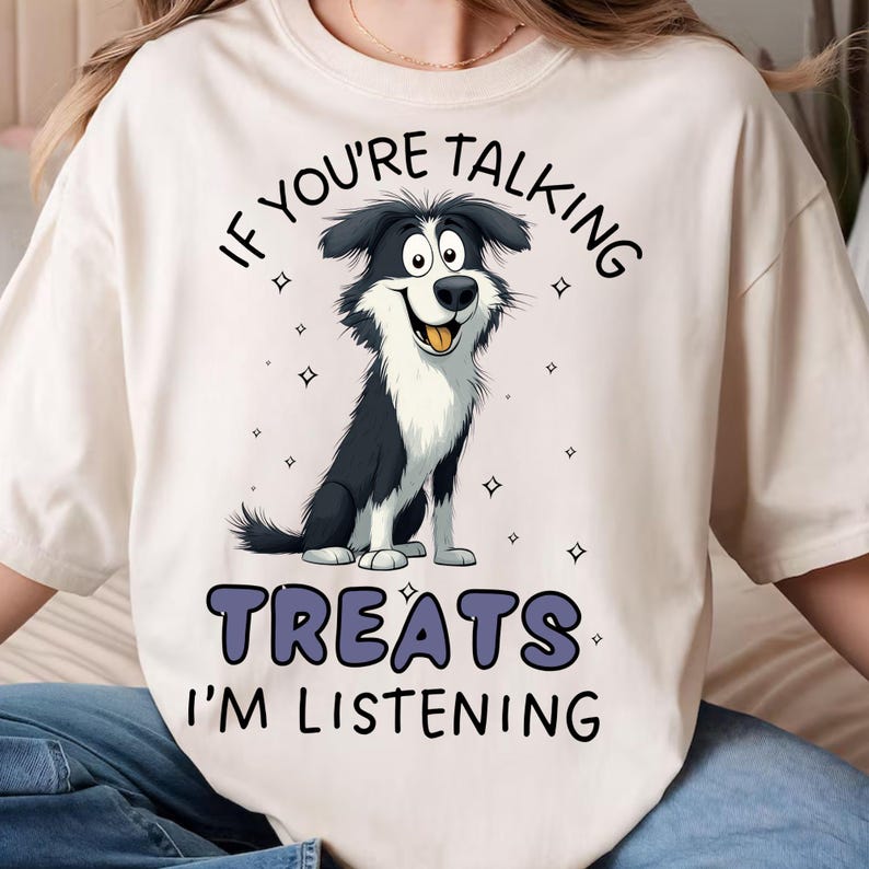 May include: Cream-colored t-shirt featuring a cartoon dog with black and white fur, a smiling expression, and its tongue sticking out. The text "IF YOU'RE TALKING TREATS I'M LISTENING" is printed above and below the dog in a dark blue color.