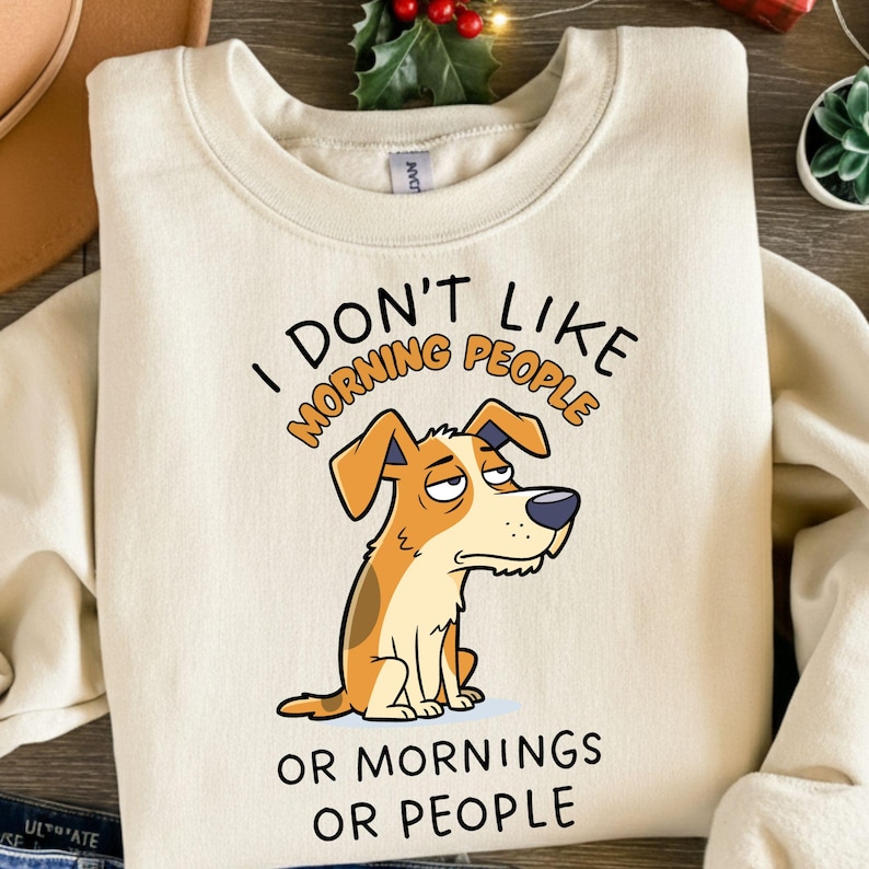 May include: Cream-colored sweatshirt with the text "I DON'T LIKE MORNING PEOPLE OR MORNINGS OR PEOPLE." A cartoon dog with a grumpy expression is featured below the text. The dog is brown and white.