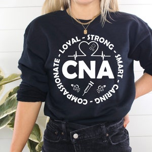 May include: Black sweatshirt with a white circle graphic that says "CNA" with a heart, EKG line, syringe, and pills around it. The text around the circle says "Caring - Smart - Strong - Loyal - Compassionate -  Advocate".