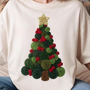 May include: Cream-colored t-shirt featuring a Christmas tree design. The tree is composed of green, red, and brown circular and star-shaped elements. A gold star adorns the top of the tree, creating a festive holiday theme.