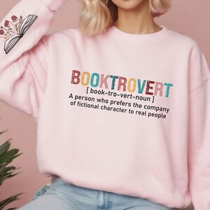 K&ouml;nnte beinhalten: Rosafarbener Pullover mit dem Text "BOOKTROVERT [book-tro-vert-noun] A person who prefers the company of fictional character to real people" auf der Vorderseite. Der Text ist in einer bunten, Retro-Schriftart.