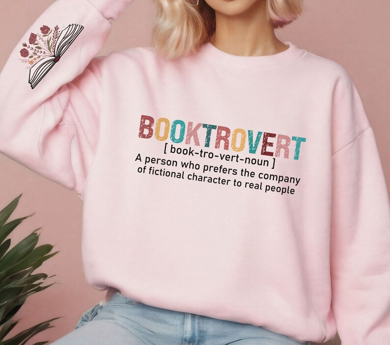 K&ouml;nnte beinhalten: Rosafarbener Pullover mit dem Text "BOOKTROVERT [book-tro-vert-noun] A person who prefers the company of fictional character to real people" auf der Vorderseite. Der Text ist in einer bunten, Retro-Schriftart.