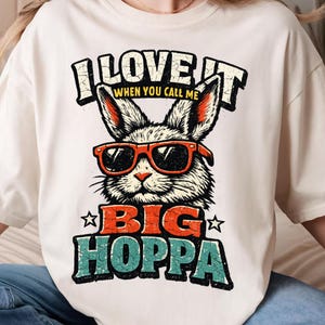 May include: Cream-colored t-shirt featuring a graphic of a rabbit wearing sunglasses. The text reads "I LOVE IT WHEN YOU CALL ME BIG HOPPA" in a distressed, vintage style. The design includes orange, teal, and black colors.