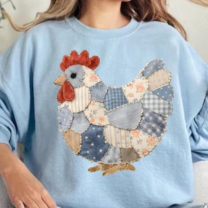 May include: A light blue sweatshirt featuring a patchwork chicken design. The chicken is made of various blue and beige fabrics with floral and striped patterns. It has a red comb and wattle, and a yellow beak and feet.