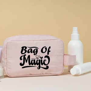 May include: A pink toiletry bag with a black inscription that reads "Bag of Magic" with a small heart design. The bag is sitting on a white surface with various white bottles and jars.