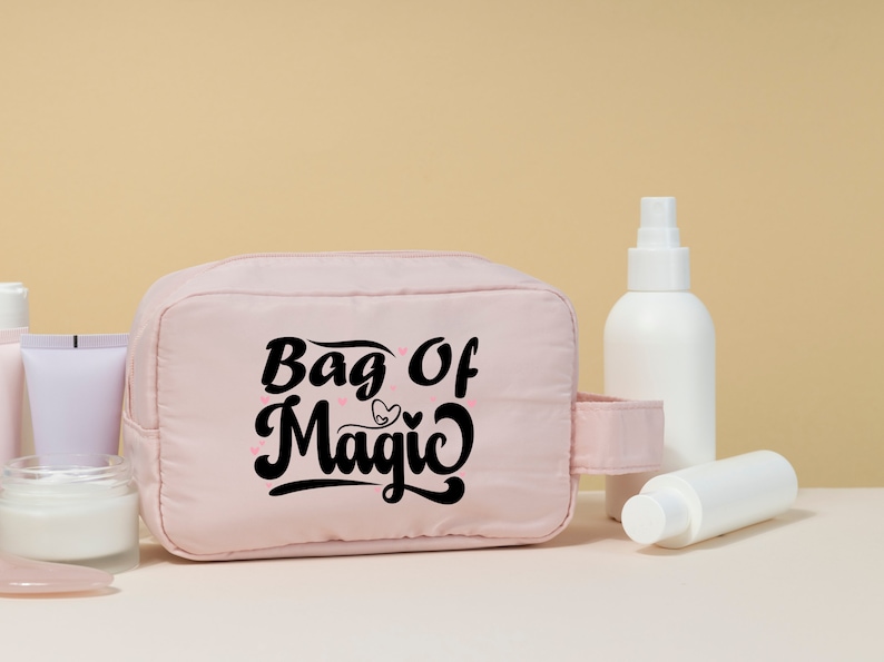May include: A pink toiletry bag with a black inscription that reads "Bag of Magic" with a small heart design. The bag is sitting on a white surface with various white bottles and jars.