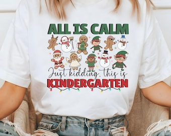 Könnte beinhalten: Weißes T-Shirt mit dem Text "ALL IS CALM Just kidding, this is KINDERGARTEN" in Grün und Rot. Das Shirt zeigt Cartoon-Illustrationen von Lebkuchenmännern, Elfen, Schneemännern und dem Weihnachtsmann, perfekt für die Weihnachtszeit.