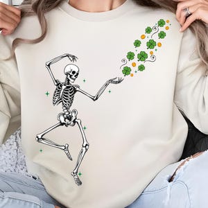 Op de afbeelding: Crèmekleurige sweatshirt met een dansende skeletafbeelding met groene klavers en oranje stippen. Het skelet staat in een speelse pose, met één been gebogen en armen uitgestrekt, alsof het de klavers vangt. Groene steraccenten zijn verspreid rond het skelet.