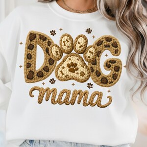 May include: White sweatshirt with a knitted design that says "Dog mama". The word "Dog" is spelled out with paw prints and a leopard print pattern. The word "mama" is in a cursive font.