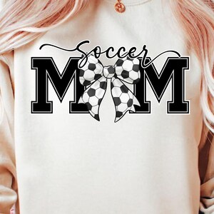May include: Cream-colored sweatshirt with the words "Soccer Mom" in black script above two large black block letter "M"s. A bow made of soccer balls is between the letters. The sweatshirt is a casual style.