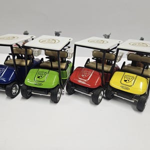 May include: Four toy golf carts in blue, green, red, and yellow. Each cart has a white roof, black frame, tan seats, and a small basket. The front of each cart displays the word "GOLF" and the words "DIE-CAST".