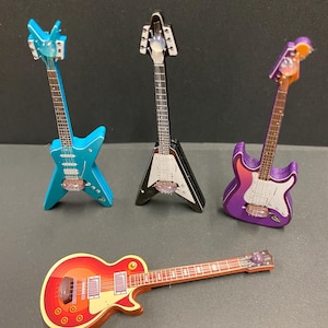 May include: Four miniature toy guitars in various shapes and colours. There is a blue star-shaped guitar, a black V-shaped guitar, a purple guitar, and a red and yellow guitar with a rounded body. Each guitar has a detailed fretboard and bridge.