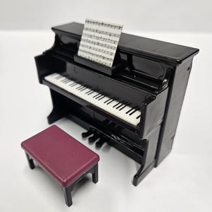 May include: A miniature black piano with white and black keys, a music stand holding sheet music, and a burgundy bench. The piano has a glossy finish and is set against a white background. The bench is rectangular with four black legs.