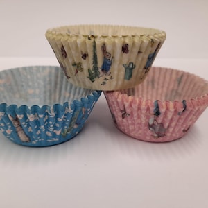 May include: Three patterned cupcake cases. One is cream with a Peter Rabbit design, one is light blue with a similar design, and the third is pink with a similar design. The cases are made of paper and have a fluted edge.