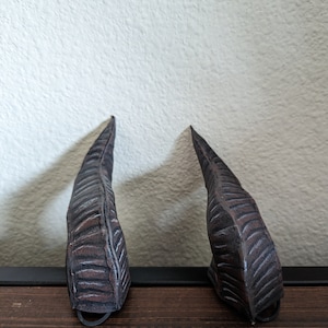 Puede incluir: Dos accesorios en forma de cuerno, de color marrón oscuro y texturados, con un anillo de metal negro en la base.