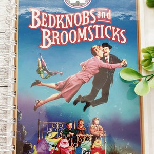 May include: A white spiral bound notebook with a colorful image of the Disney movie Bedknobs and Broomsticks. The cover features the title of the movie in red and white letters, and a scene from the movie with a couple dancing in the air.
